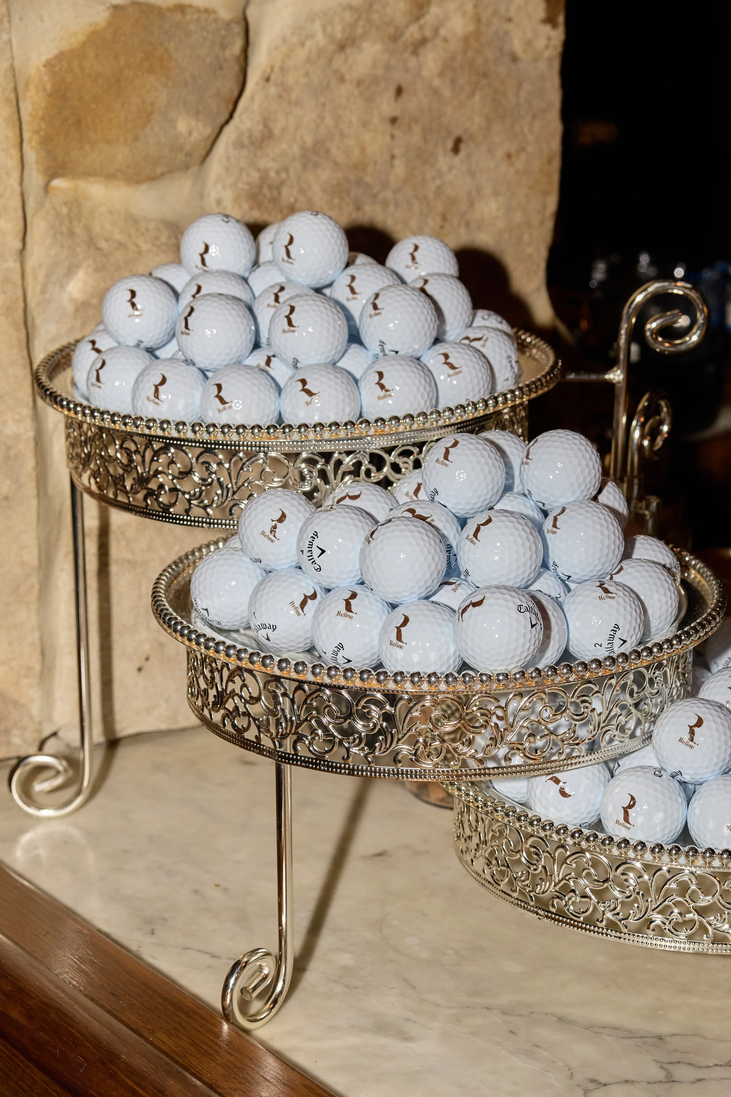 Two-tiered metal stand filled with white golf balls, each marked with a brown logo, placed on a marble surface against a stone wall background.