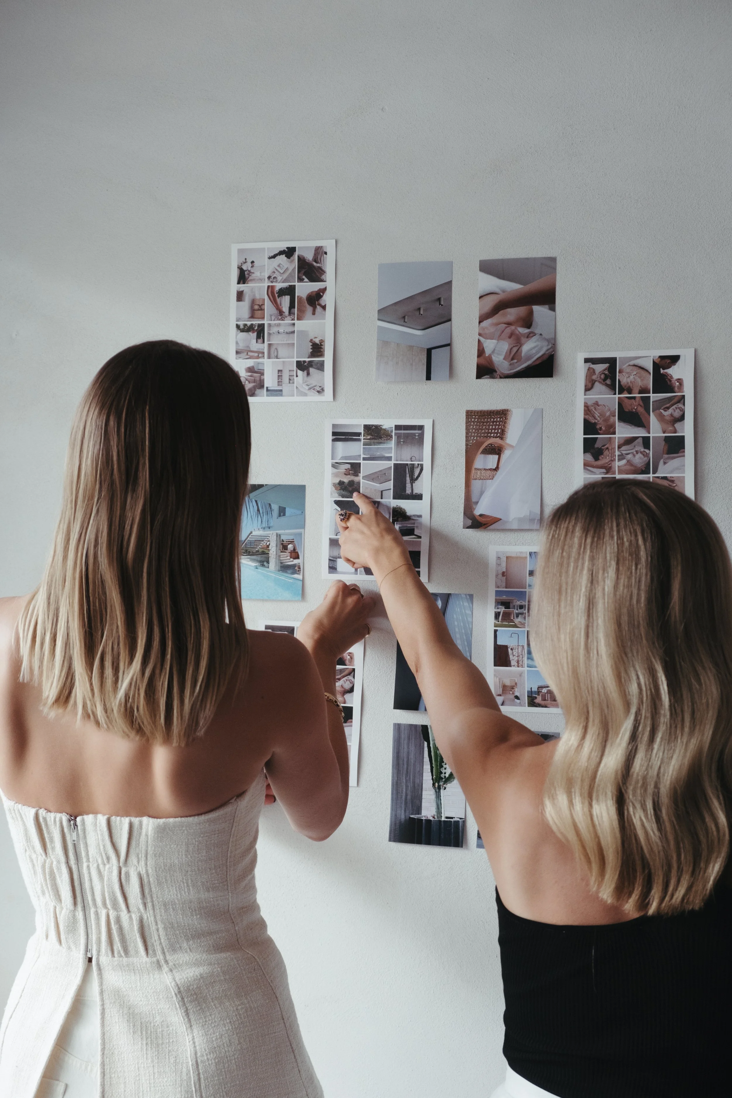 Two women examining a wall with various photos and mood board images for interior design or decorating.