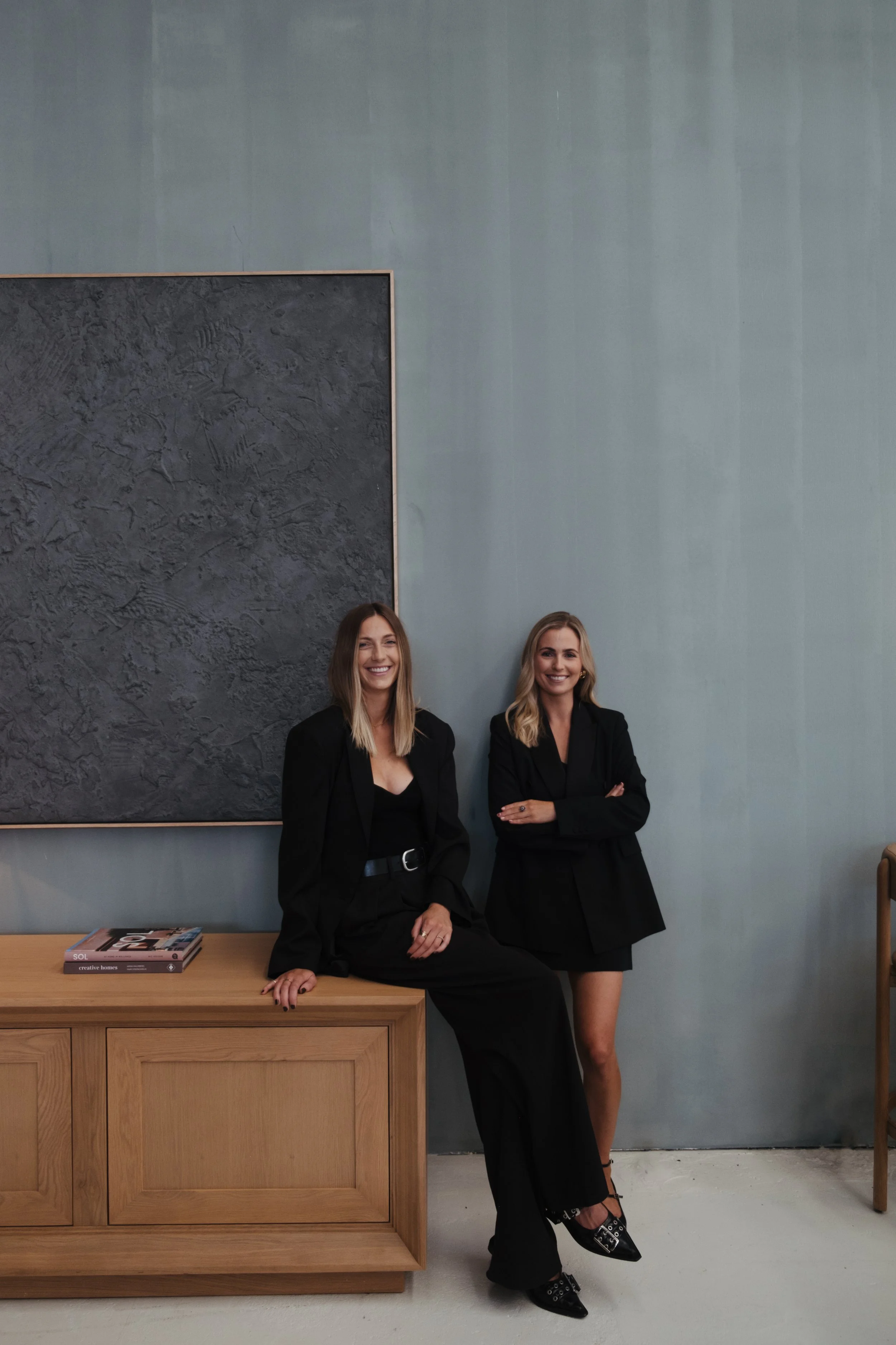 Two women in black outfits smiling and posing indoors; one is sitting on a wooden cabinet, the other is standing with arms crossed against a minimalist gray wall with a textured black framed artwork.