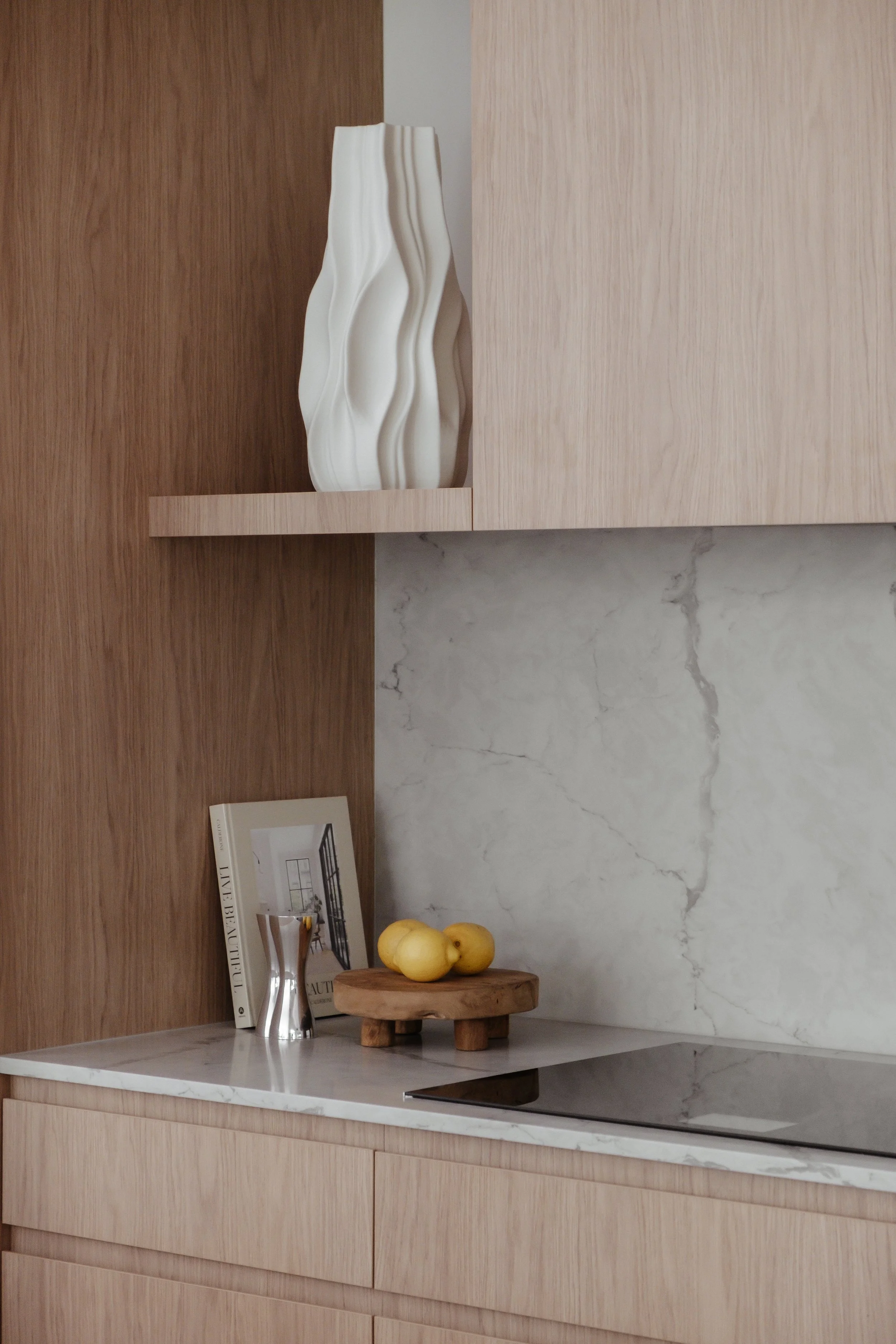 A kitchen countertop with a small cutting board holding two yellow lemons, a magazine, and a silver pitcher, with a marble backsplash and wooden cabinetry.