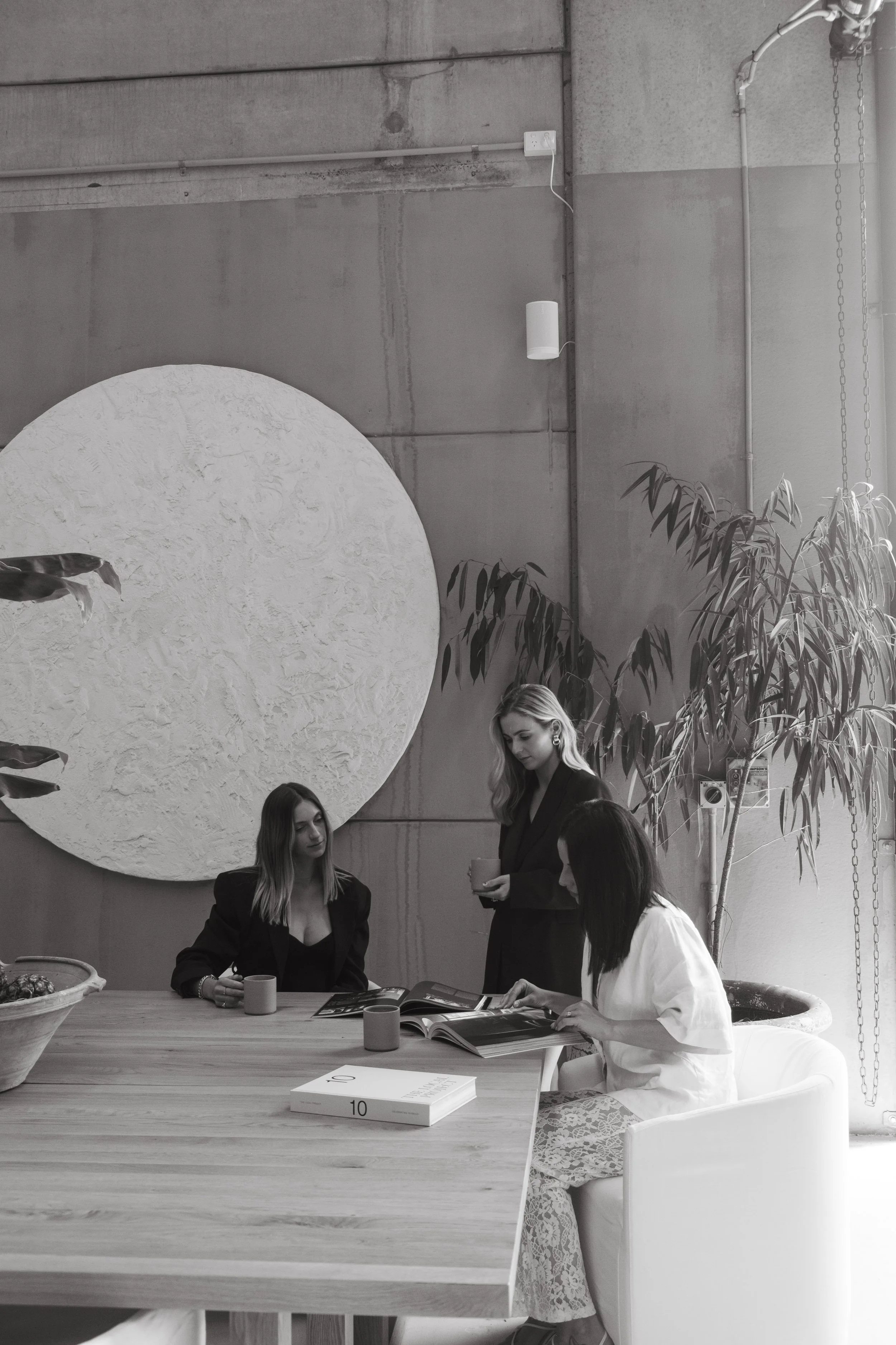 Black and white photo of three women in a modern, industrial-style room with a large textured circular wall art piece, a tall plant, and a wooden table with magazines and cups.