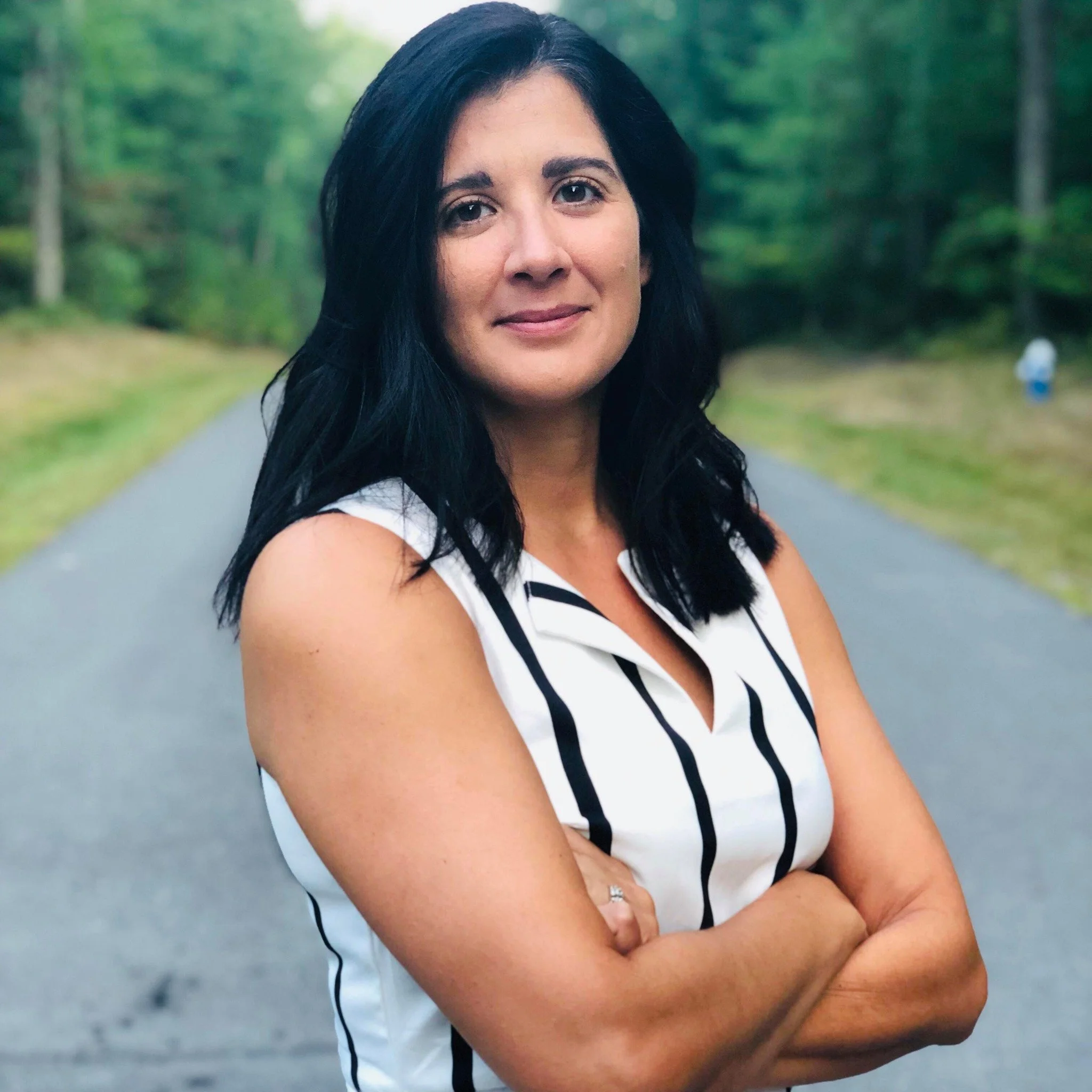 Person with dark hair wearing a sleeveless, striped shirt, standing on a road surrounded by trees, arms crossed.