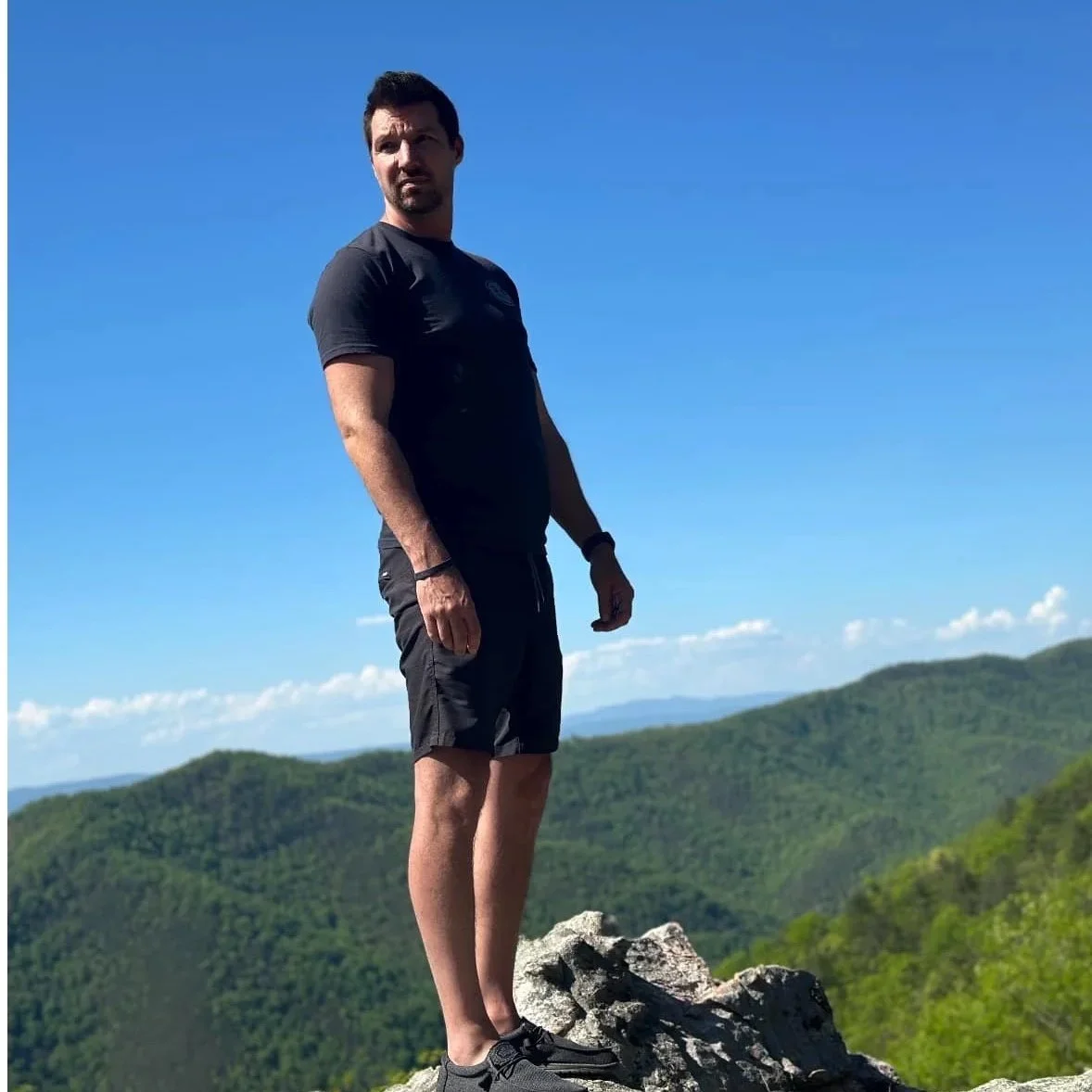Person in blue shirt standing outside with greenery in the background