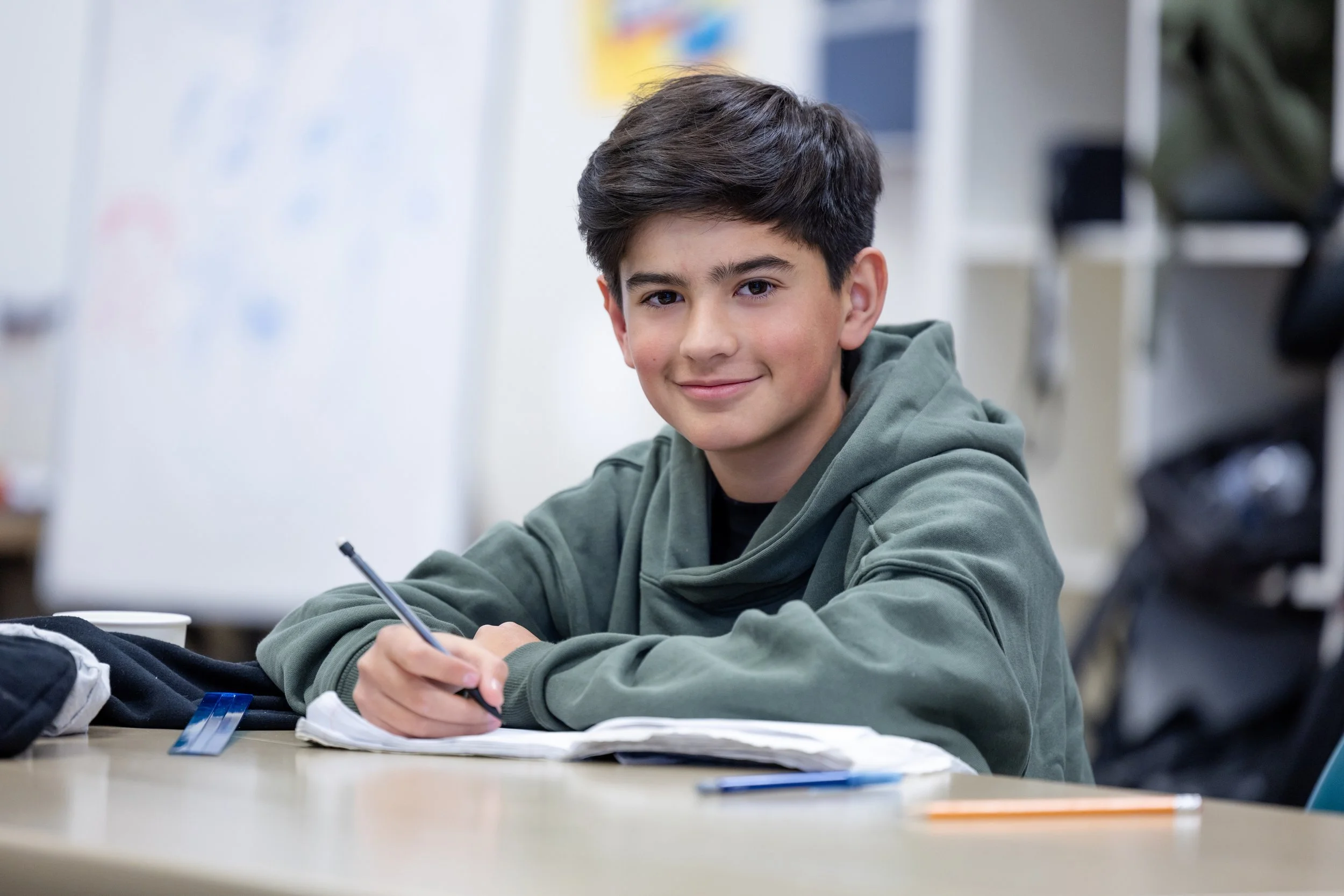 A boy wearing a green hoodie smiles at the camera while writing in a notebook during an activity at the Boys & Girls Clubs of Monterey County.