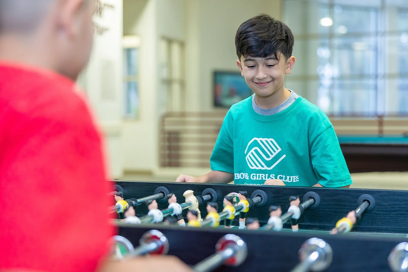 A boy wearing a green Boys & Girls Clubs of Monterey County T-shirt plays foosball with a friend, smiling as he focuses on the game inside the Club’s recreation area.