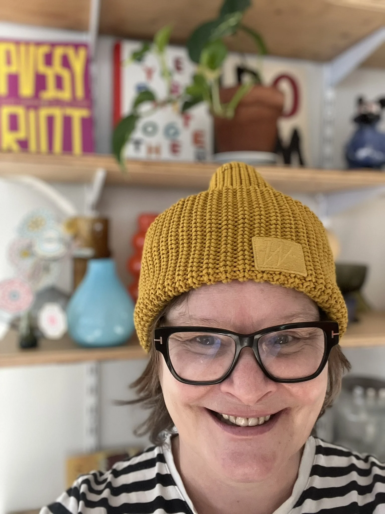 Ihminen hymyilee ja otti selfietä sisätiloissa, taustalla hylly täynnä erilaisia astioita ja koriste-esineitä.