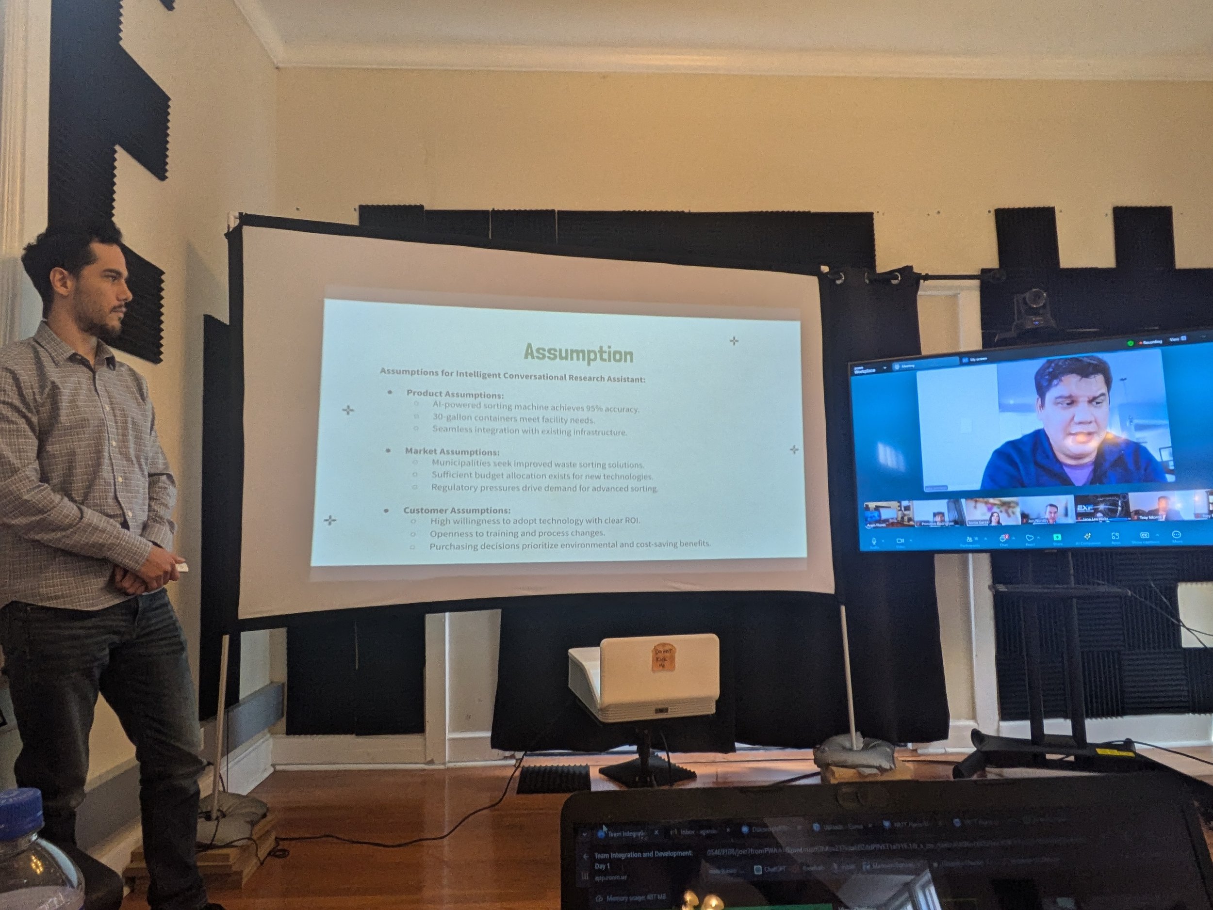 Man presenting in front of a projection screen with presentation slides, with a large monitor displaying a video call with multiple participants.