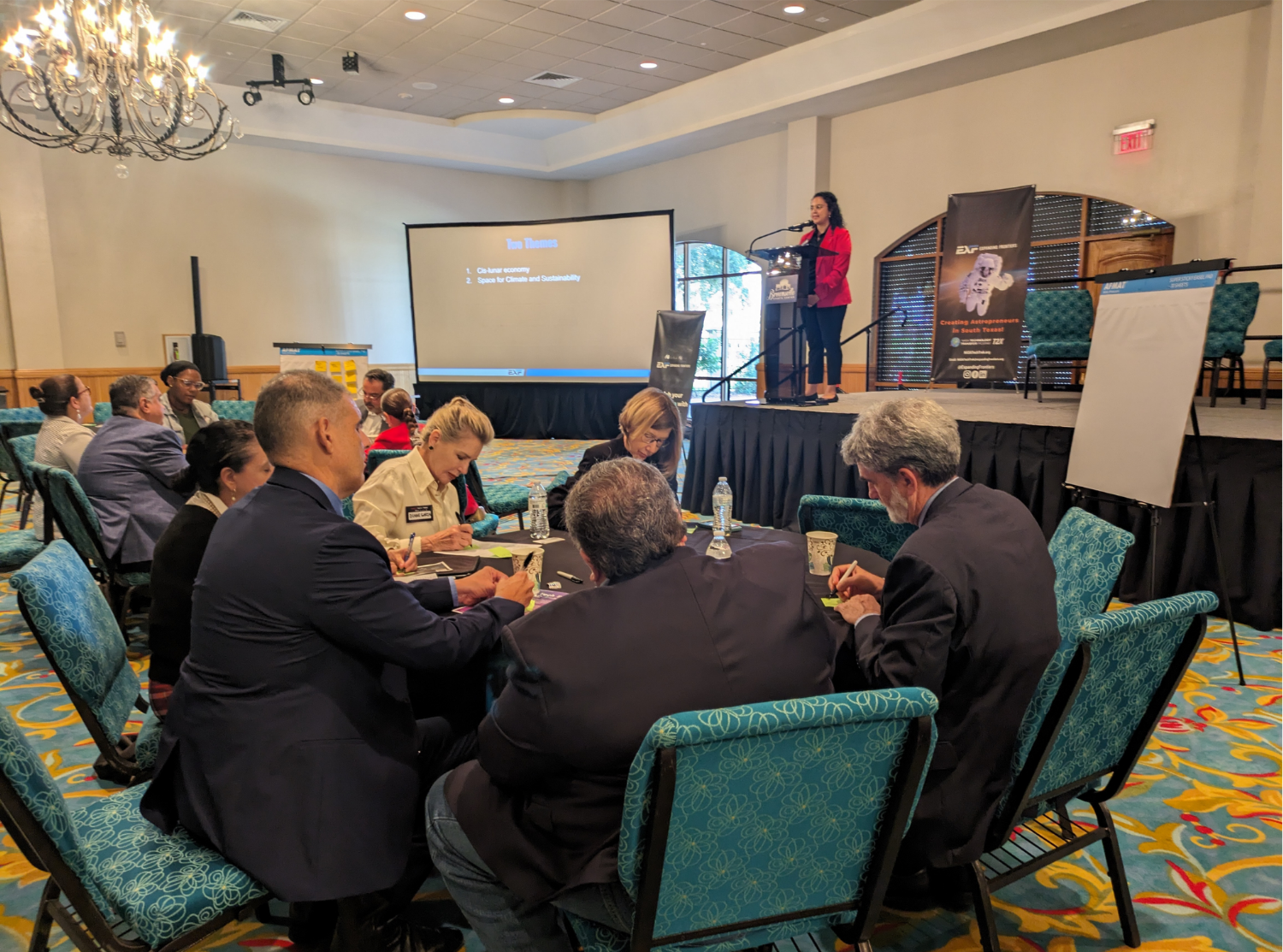 A woman giving a presentation at a conference, standing on a stage with a podium, large screen displaying a slide, and audience seated at tables with notebooks and water bottles.