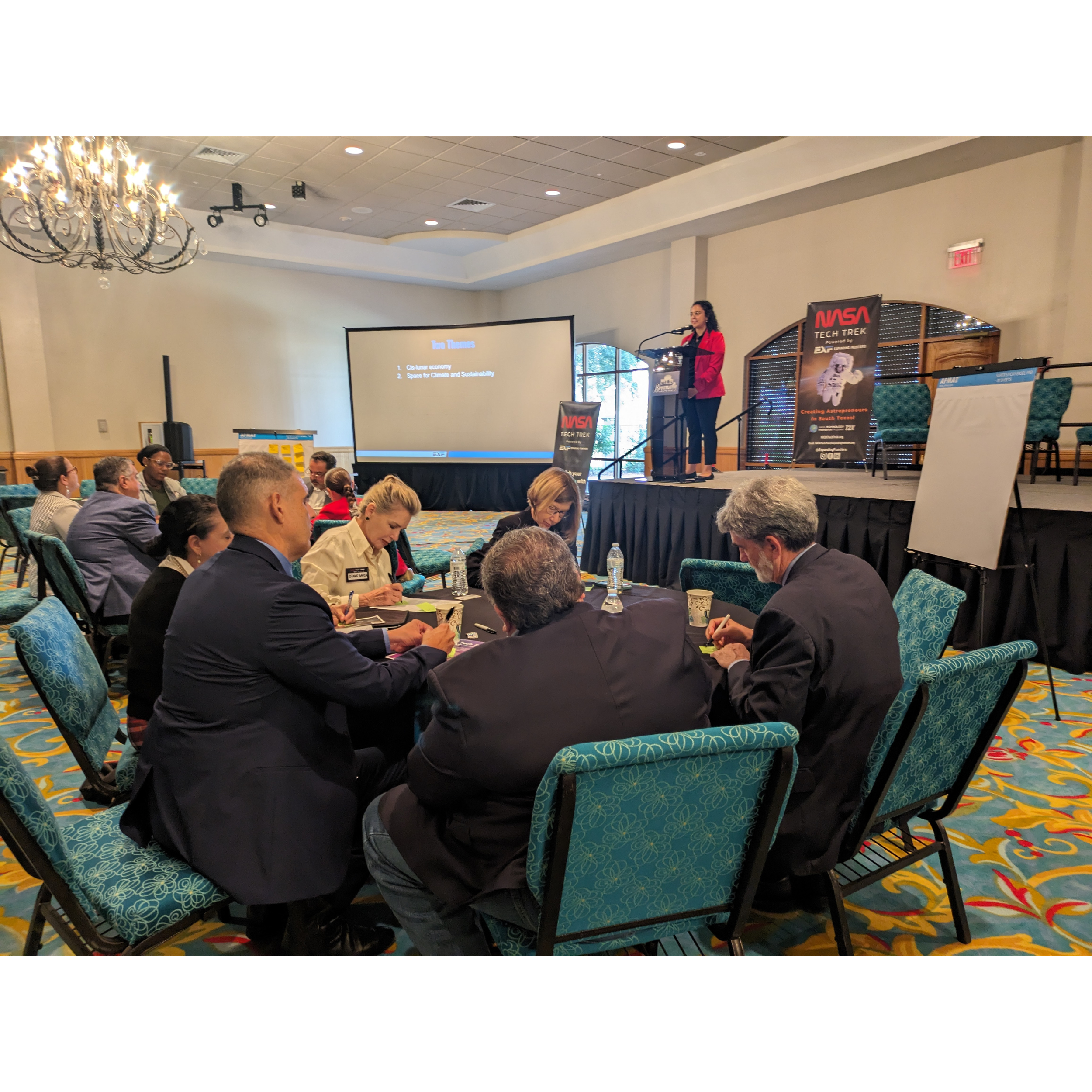 A woman gives a presentation on a stage at a conference, with a large screen behind her displaying text, while an audience listens and takes notes at tables.