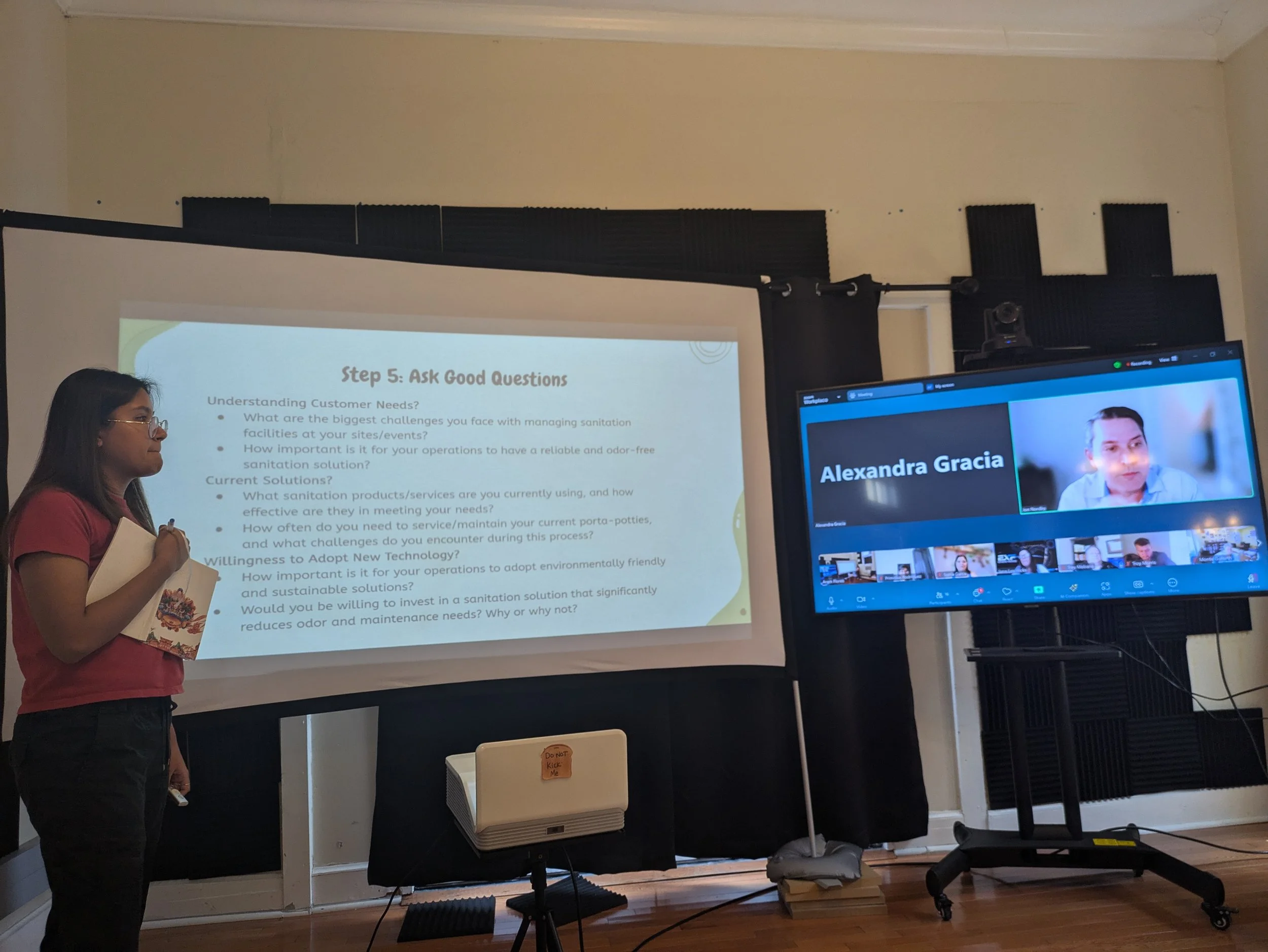 A woman giving a presentation in front of a screen with slides, and a woman participating via video call on a large monitor.