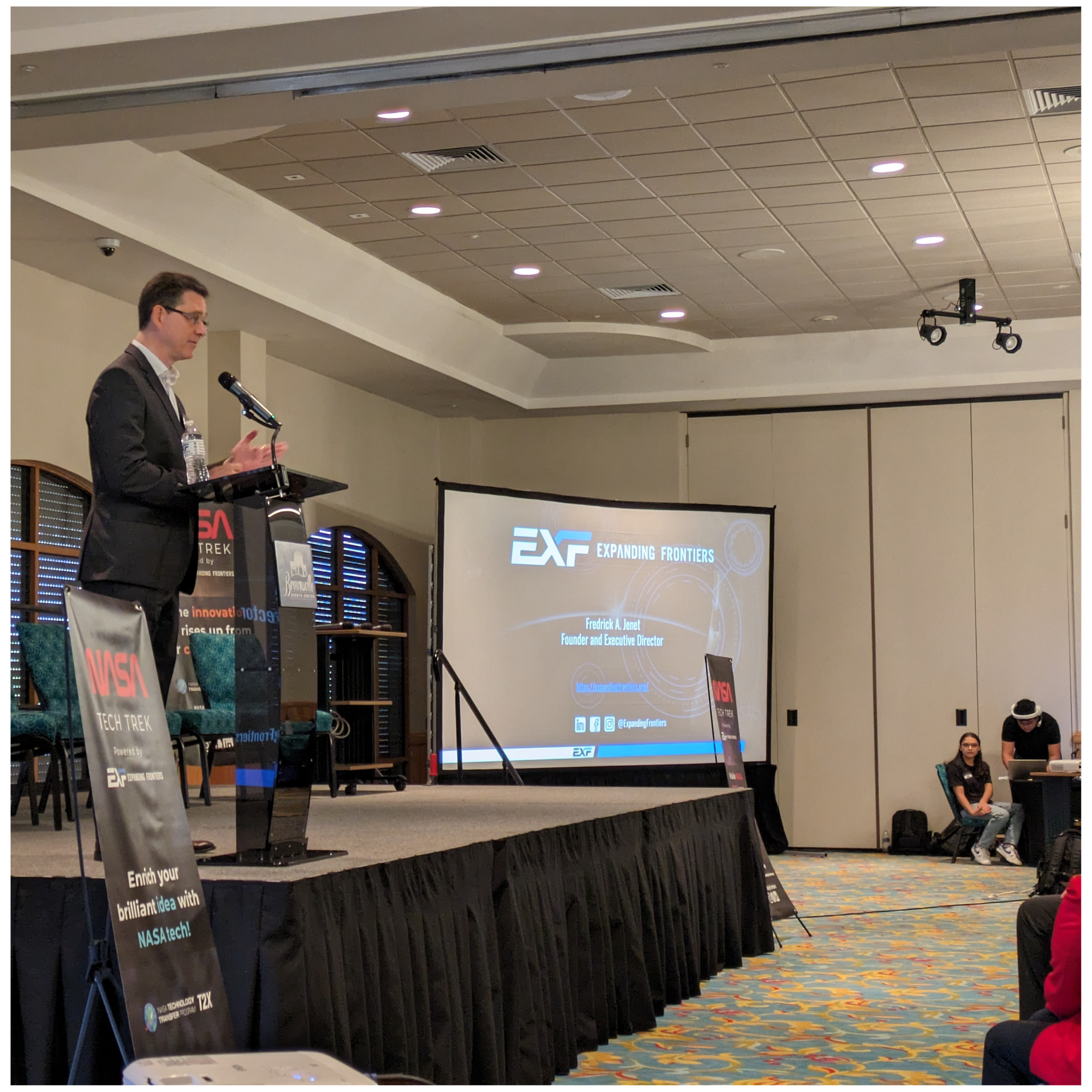 A man in a black suit speaking at a podium on a stage with a large screen behind him displaying a presentation titled 'Expanding Frontiers' by Fredrick A. Junet, Founder and Executive Director. The audience is seated on the right side, with some members visible.