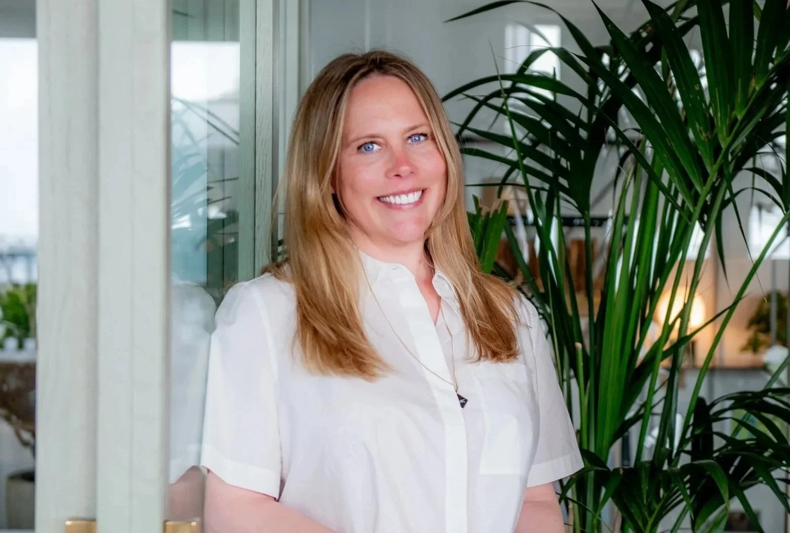 Founder, Gayle Jones, Tide & Bloom smiling to camera wearing a white shirt standing next to green plant