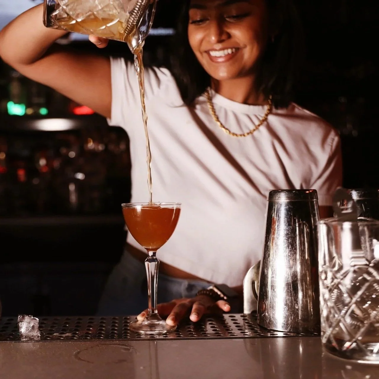 A woman smiling as she pours a drink into a cocktail glass on a bar counter, with bar tools including a shaker and glasses nearby.