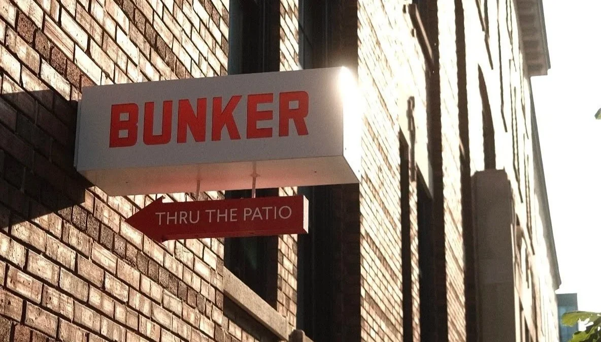 A sign with the word 'BUNKER' in red letters over a brick building, with an arrow pointing to the left that reads 'THRU THE PATIO'.