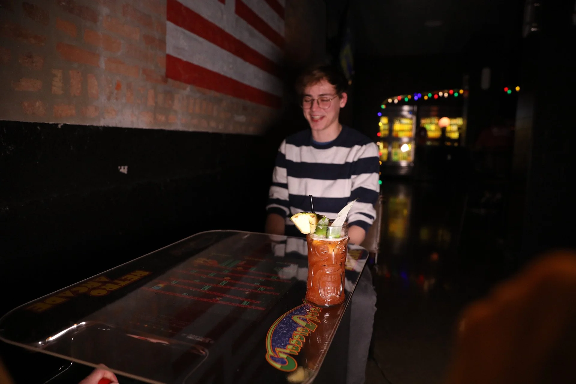 A young man in glasses and a striped shirt sitting at a bar, with a colorful tiki-style drink in front of him on the table.