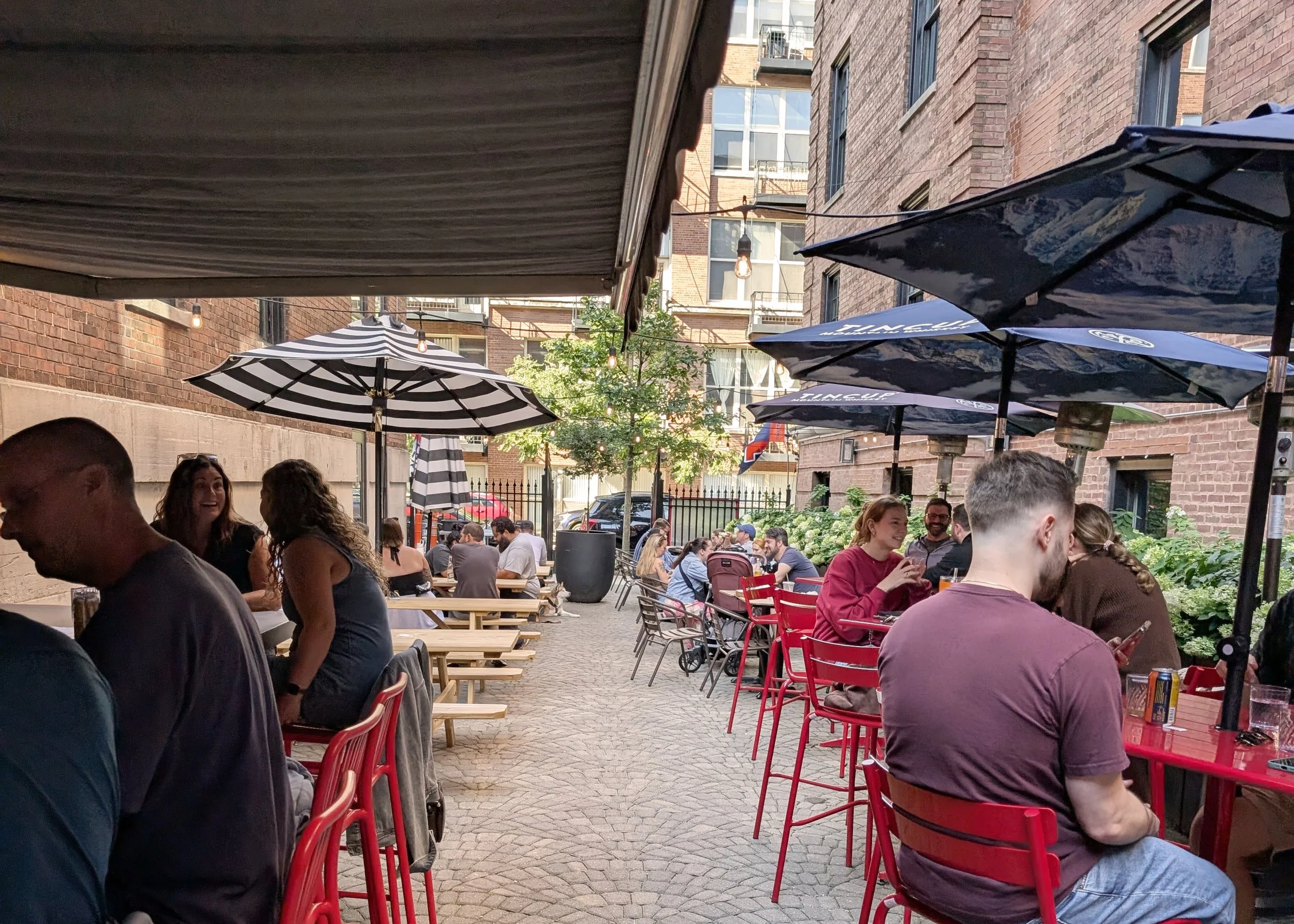 Outdoor patio with people dining at tables, some using umbrellas, surrounded by brick buildings and greenery.