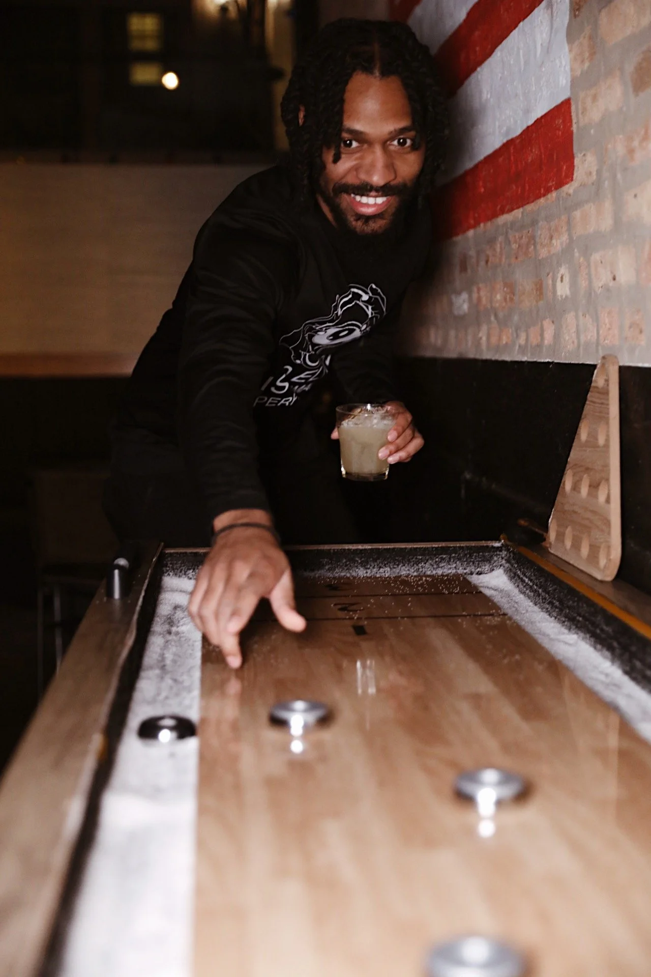 Man with dreadlocks smiling and serving drinks at a shuffleboard table in a dimly lit bar or arcade.