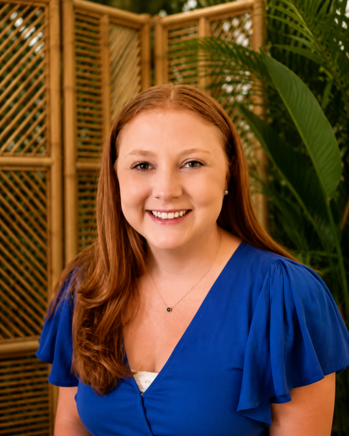 A young woman with long red hair, smiling, wearing a blue top with puffed sleeves and a delicate necklace.