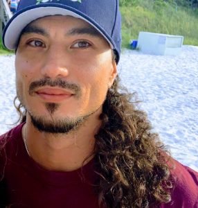 Portrait of a man with long curly hair, wearing a blue cap and a maroon shirt, outdoors near a beach with sand and trees in the background.