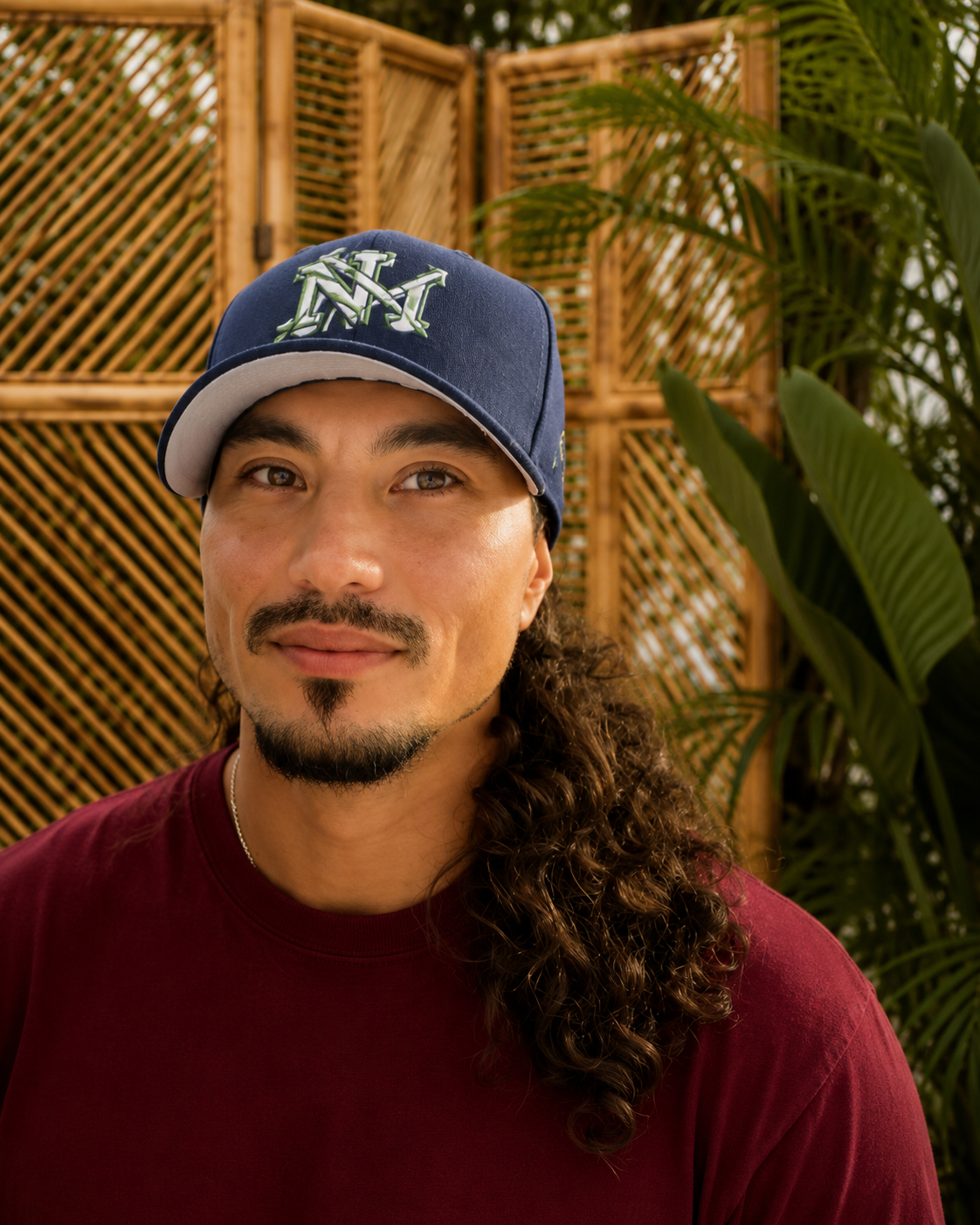 Portrait of a man with long curly hair, wearing a blue cap and a maroon shirt, outdoors near a beach with sand and trees in the background.