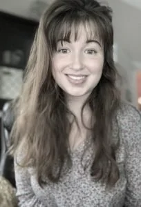A young woman with long, wavy brown hair and bangs, smiling at the camera, wearing a patterned top indoors.