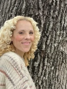 A woman with curly blonde hair smiling next to a large tree with textured bark.