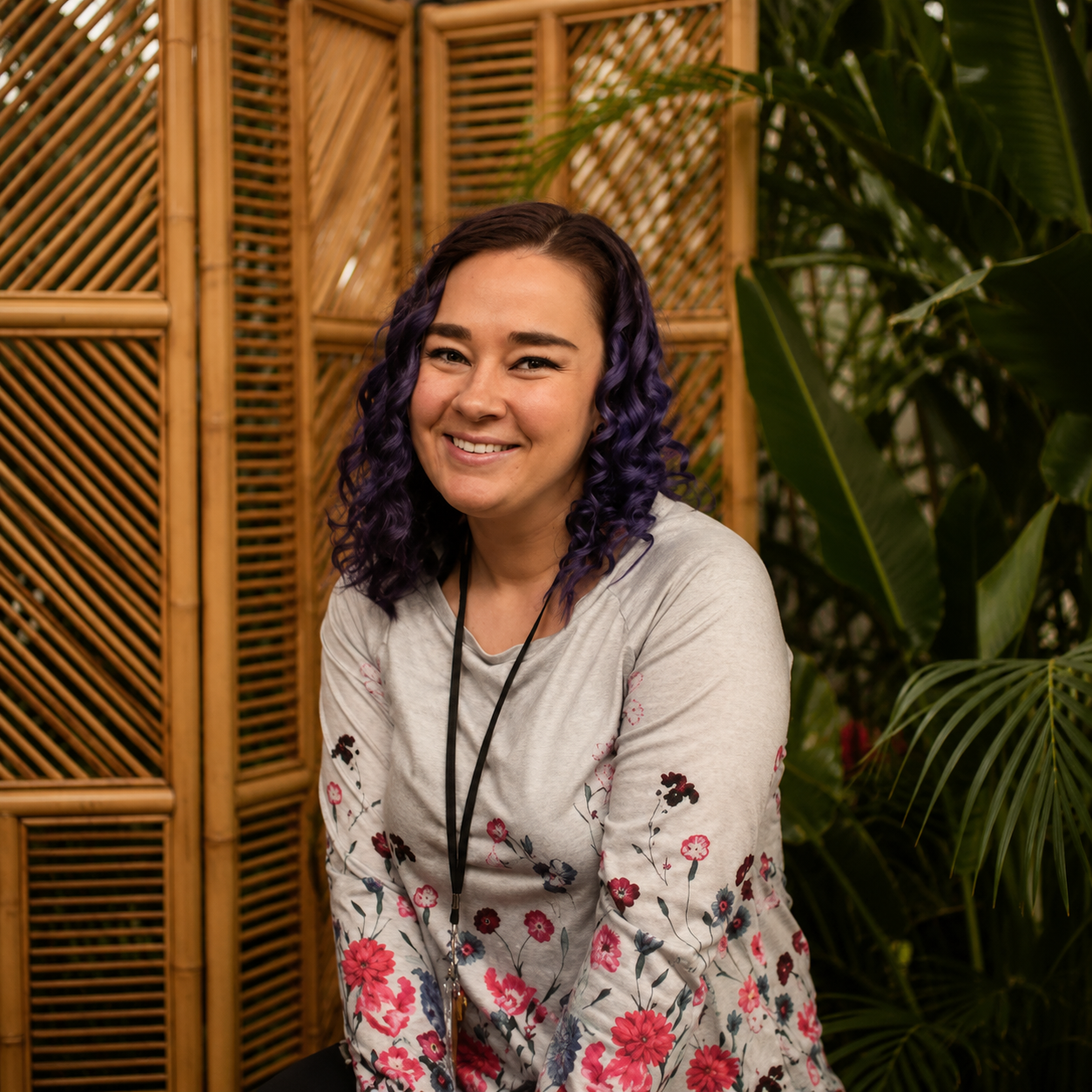 Young woman with purple curly hair smiling in front of a brick wall decorated with red flowers.