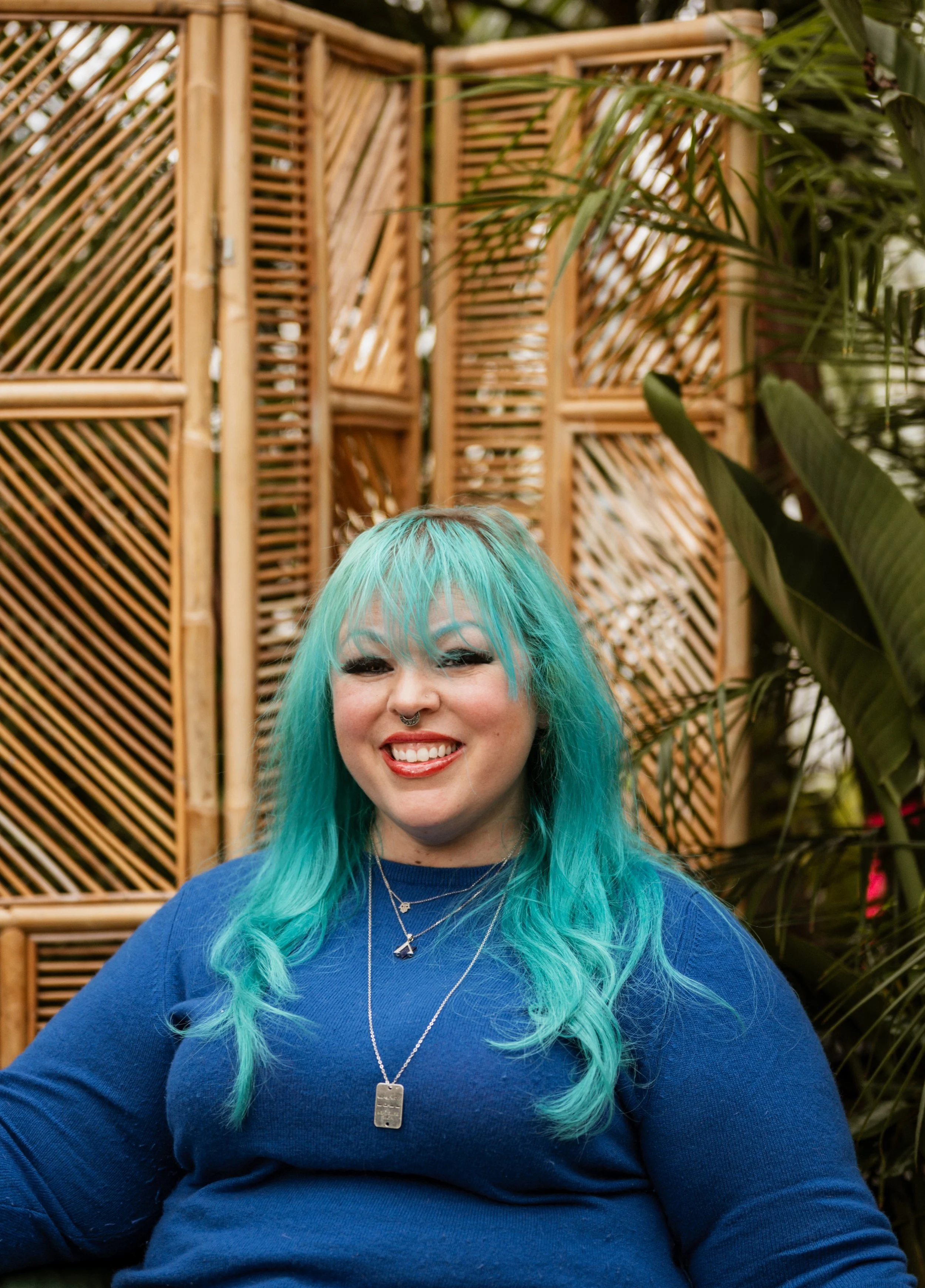 A woman with bright turquoise hair, wearing a blue top, multiple necklaces, and smiling, standing in front of a wooden lattice divider with green plants around her.