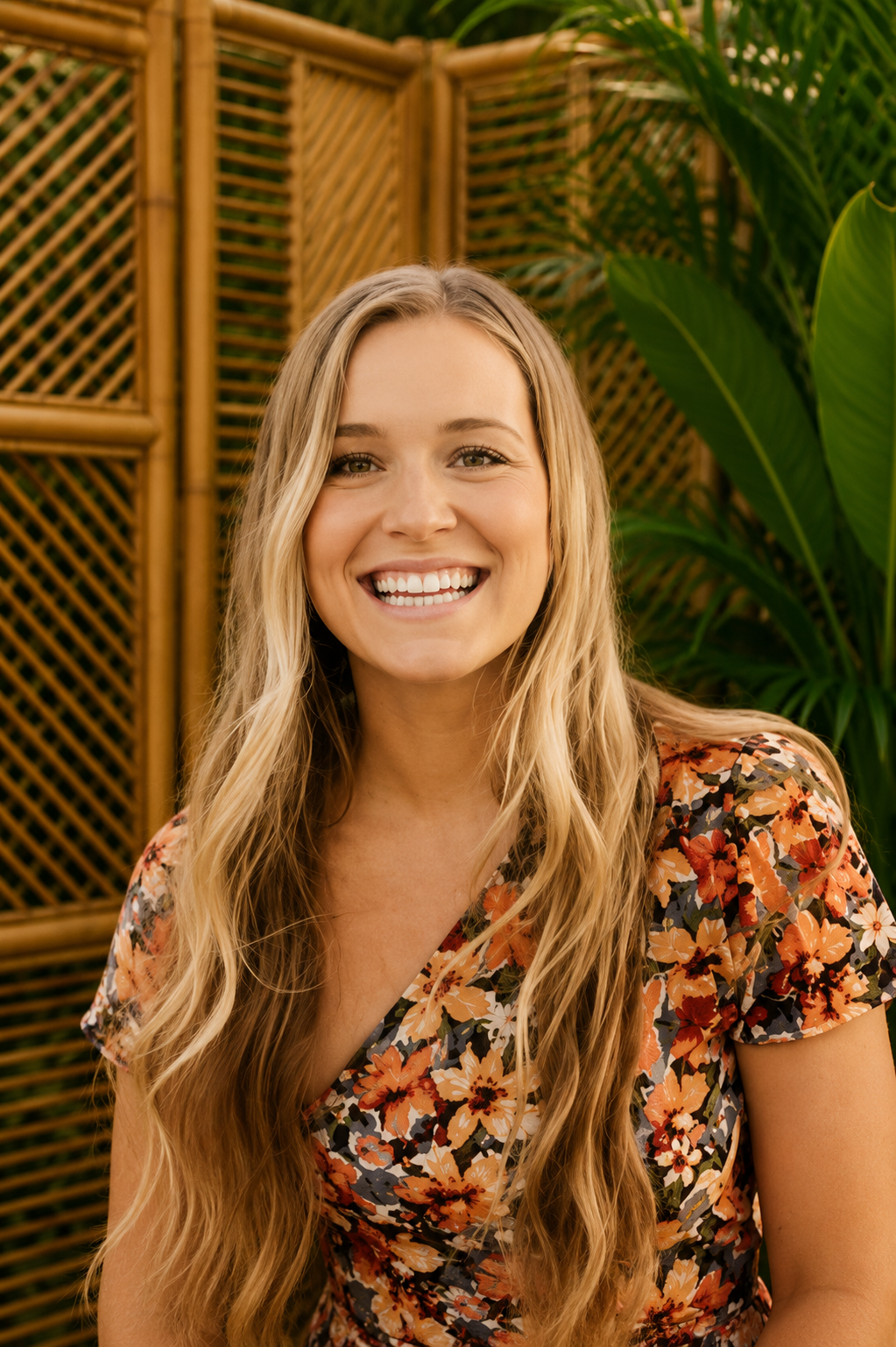 A young woman with long blonde hair smiling at the camera, wearing a floral dress, outdoors with a cloudy sky in the background.