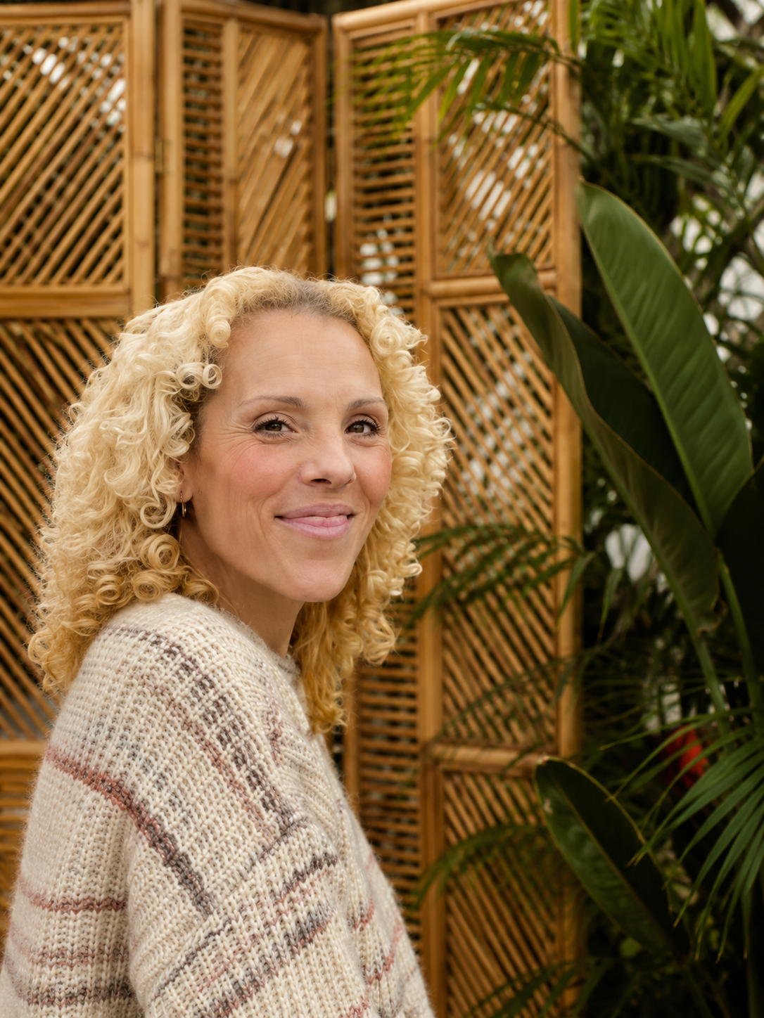 A woman with curly blonde hair smiling next to a large tree with textured bark.