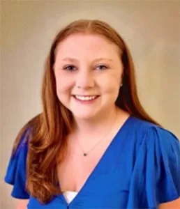 A young woman with long red hair, smiling, wearing a blue top with puffed sleeves and a delicate necklace.