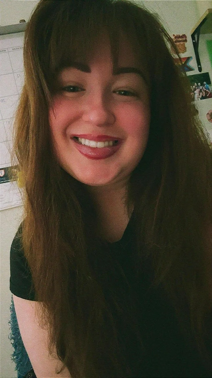 A young woman with long, wavy brown hair and bangs, smiling at the camera, wearing a patterned top indoors.