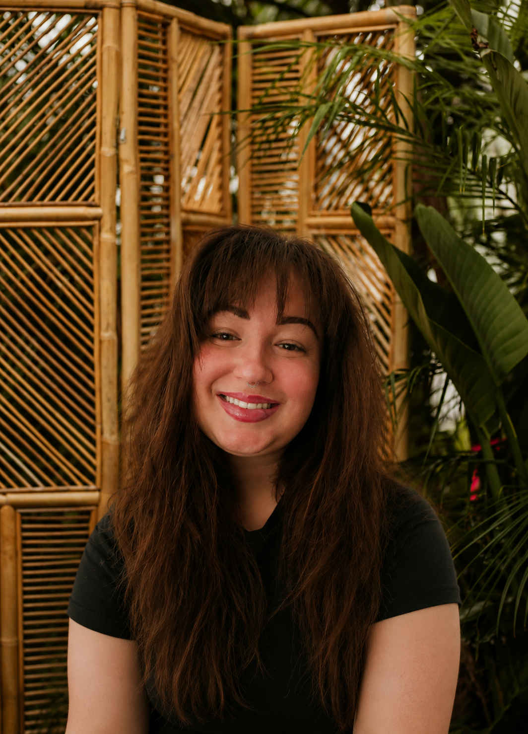 A young woman with long, wavy brown hair and bangs, smiling at the camera, wearing a patterned top indoors.