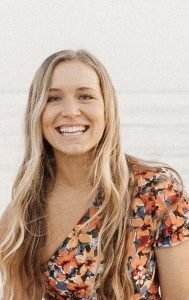 A young woman with long blonde hair smiling at the camera, wearing a floral dress, outdoors with a cloudy sky in the background.