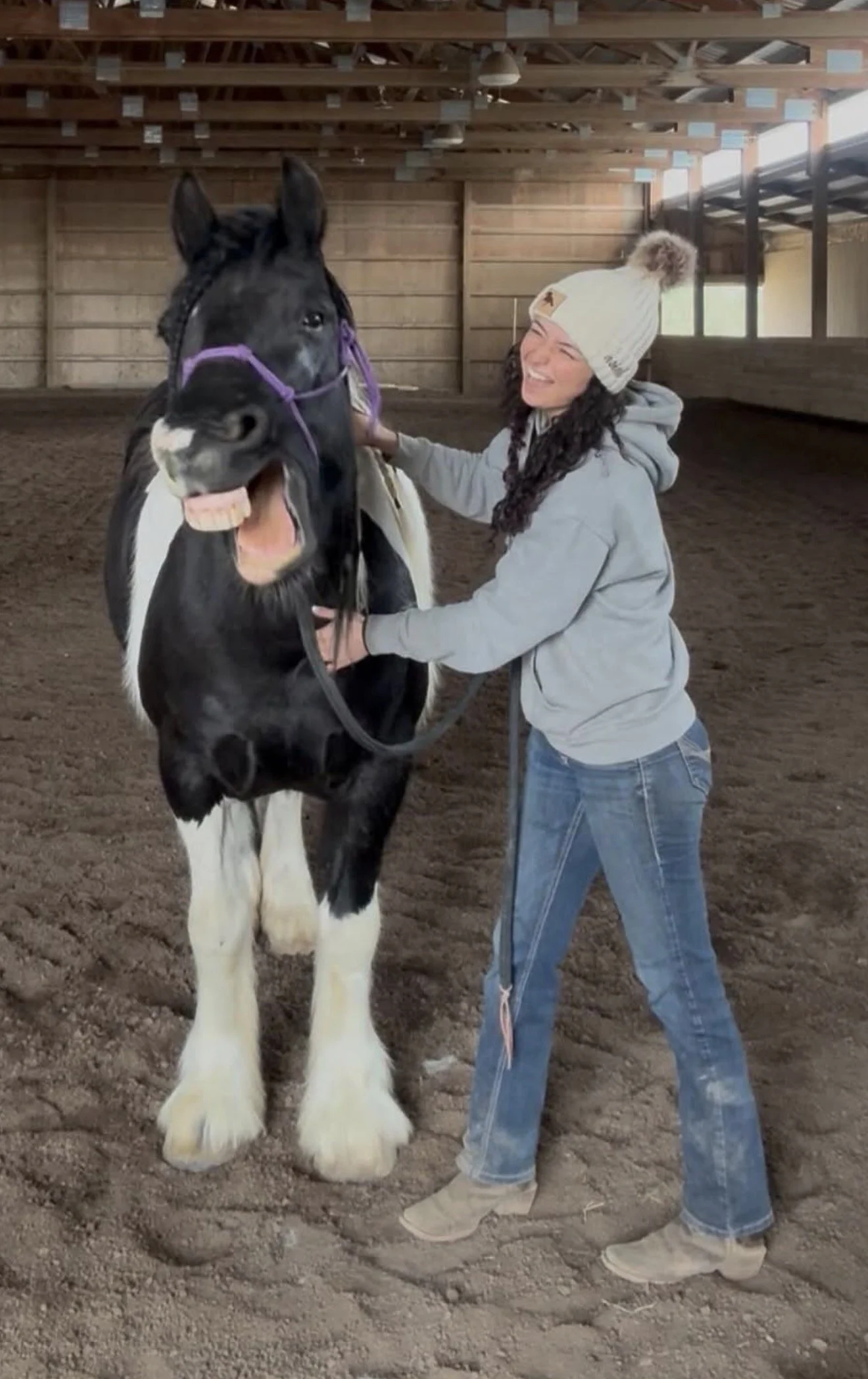 A woman smiling in a grey hoodie and blue jeans, wearing a white knit hat, standing inside an indoor riding arena holding a black and white horse's bridle, with the horse showing a playful expression with its mouth open.