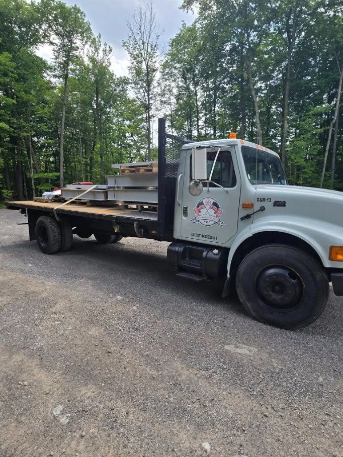 White utility truck with a logo on the door, parked on a gravel area with trees in the background, carrying ladders, wood, and construction materials.