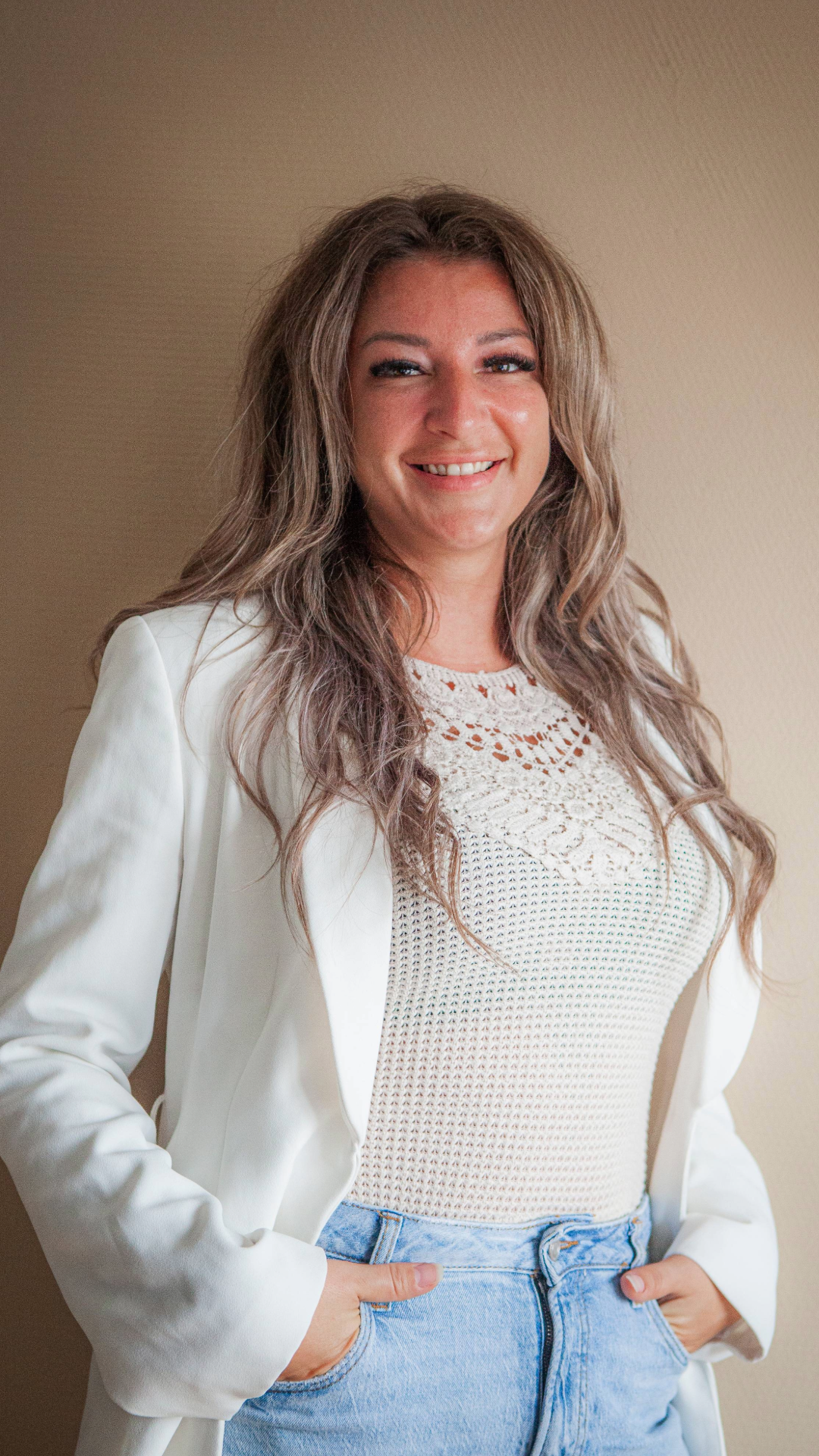 Vrouw met lange, gekrulde haar in een witte blazer en trui, poseert lachend voor een beige muur.