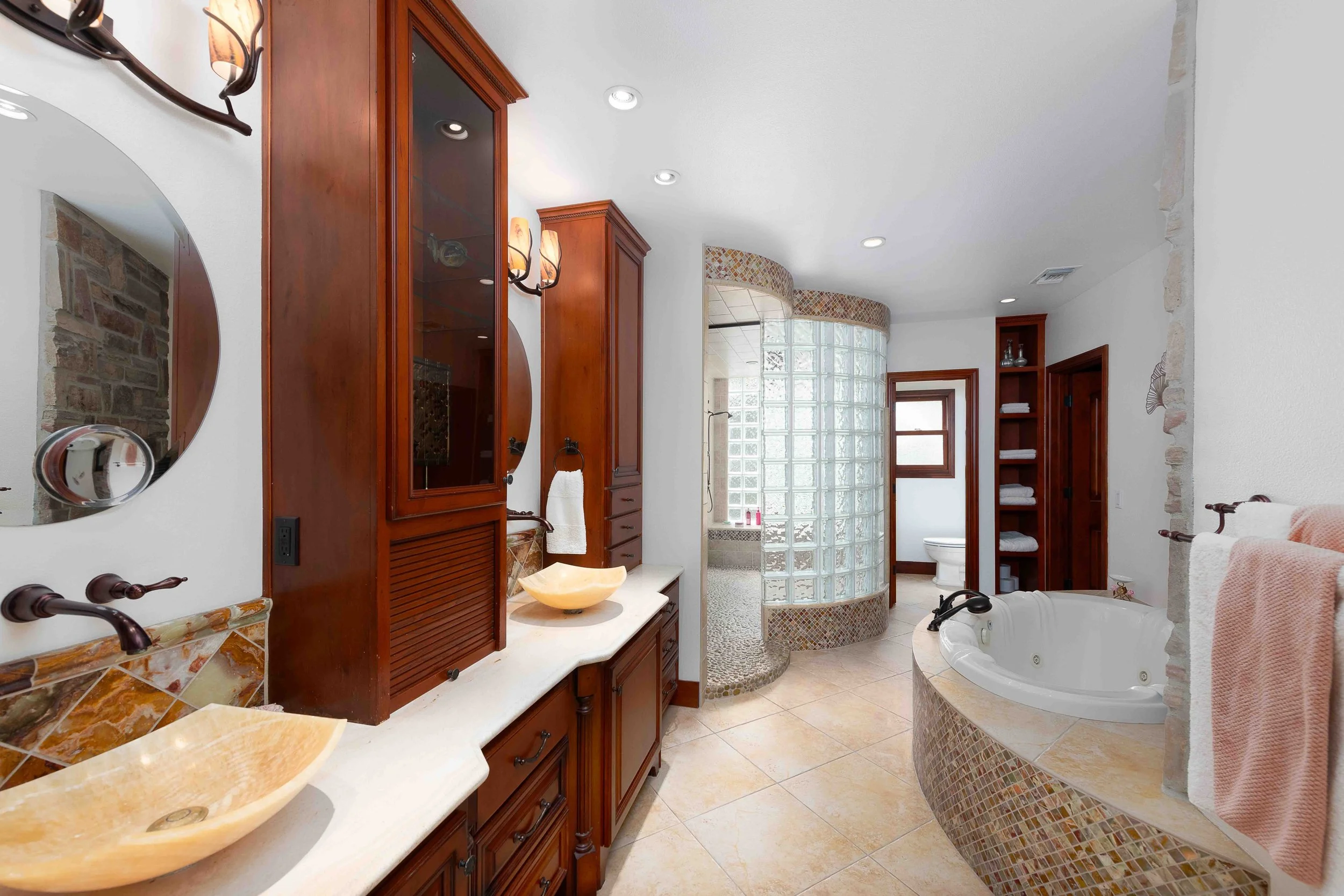 A spacious bathroom with dual sinks, wooden cabinetry, a glass block shower, a bathtub with mosaic tiles, and built-in shelves with towels.