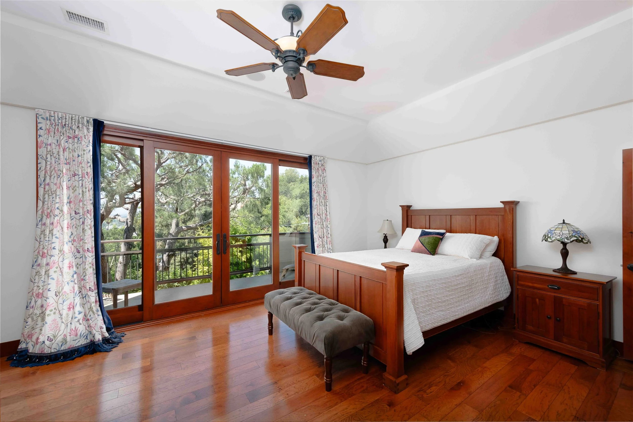Bedroom with wooden furniture, large sliding glass doors leading to a balcony, trees outside, hardwood floor, white bedspread, and floral curtains.
