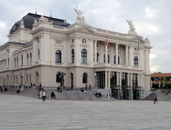 A grand, historic white building with classical architecture, topped with statues, large windows, columns, and flags. People are sitting on the steps in front.