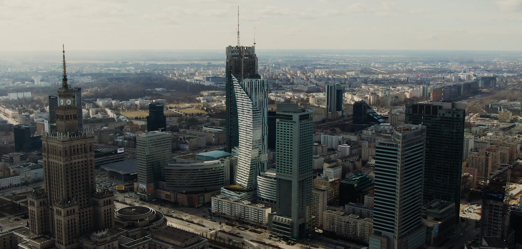 Aerial view of a city skyline featuring tall skyscrapers, including some with modern glass designs, with a mix of older and new buildings extending into the distance under a cloudy sky.