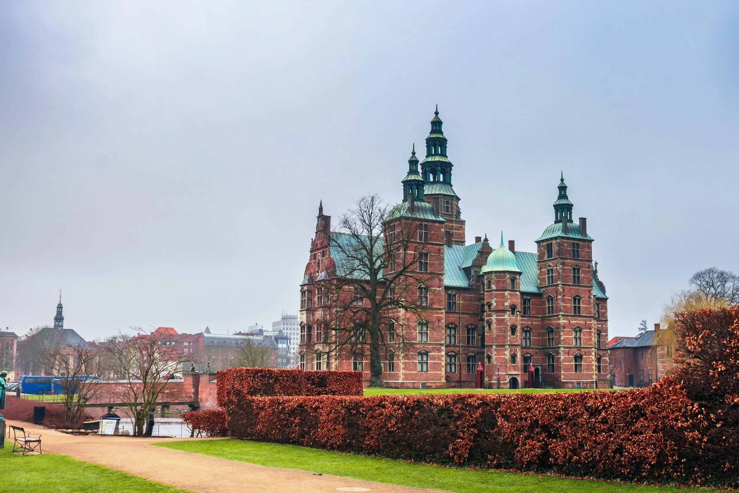 A historic castle with multiple turrets and green domed roofs, surrounded by landscaped gardens and trees, under a cloudy sky.