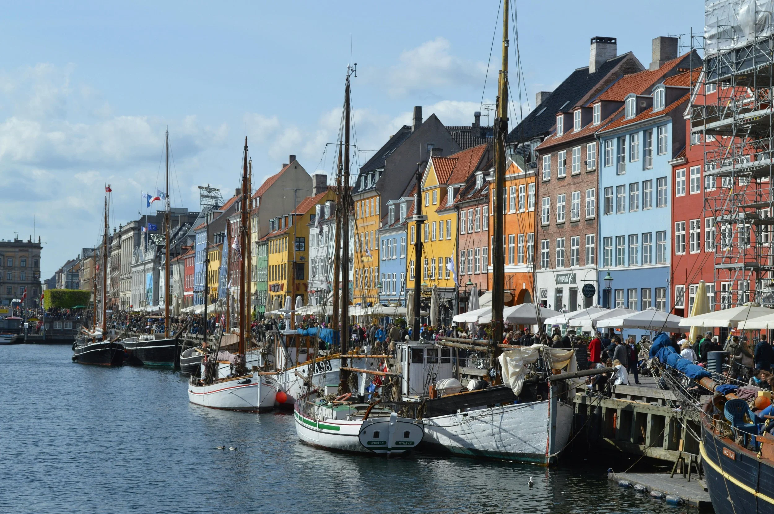 Colorful row of houses along a canal with several sailboats docked in front and people walking along the waterfront.