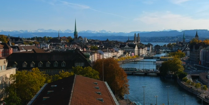 A cityscape of Zurich, Switzerland, featuring the Limmat River, historic buildings, church spires, and snow-capped mountains in the distance under a clear sky.