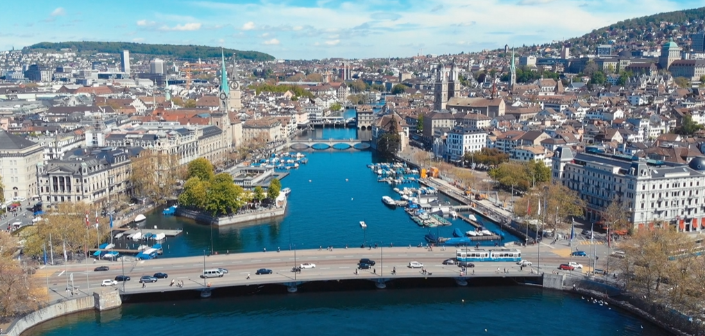 Aerial view of a city with a river running through the center, boats docked along the river, a bridge crossing the river, and historic buildings lining the streets. Hills with buildings rise in the background under a partly cloudy sky.