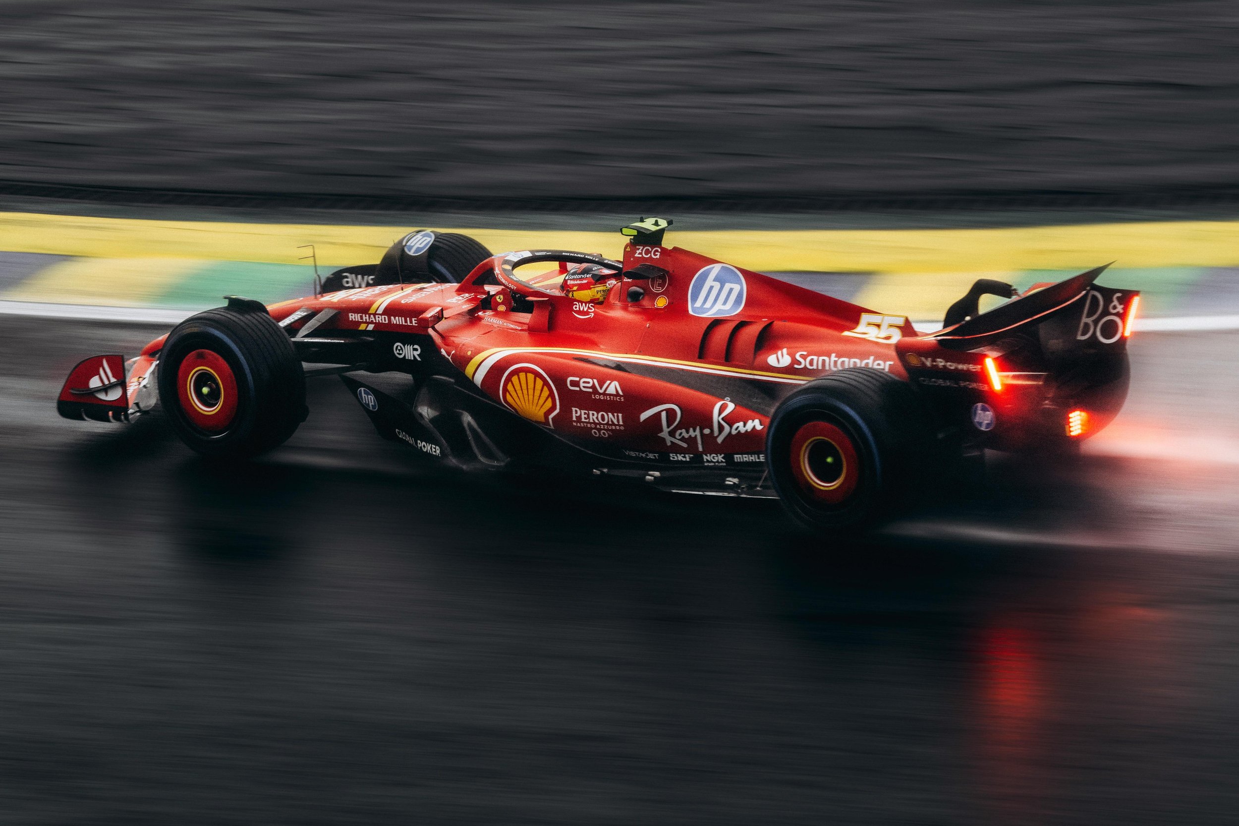 Red Formula 1 race car speeding on wet track with blurred background, rain reflected on road surface, and rear lights on.