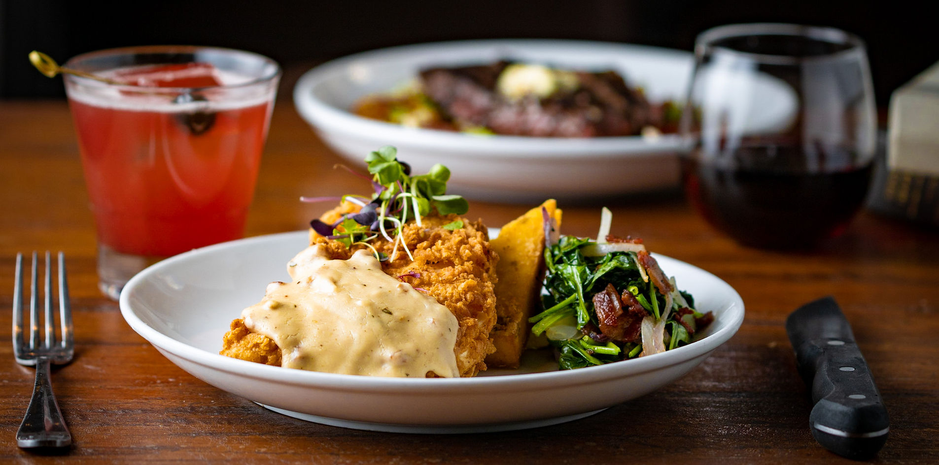 Plate of crispy fried chicken with gravy, accompanied by sautéed greens and a wedge of cornbread. A cocktail with a dark cherry and a wine glass are in the background on a wooden table.