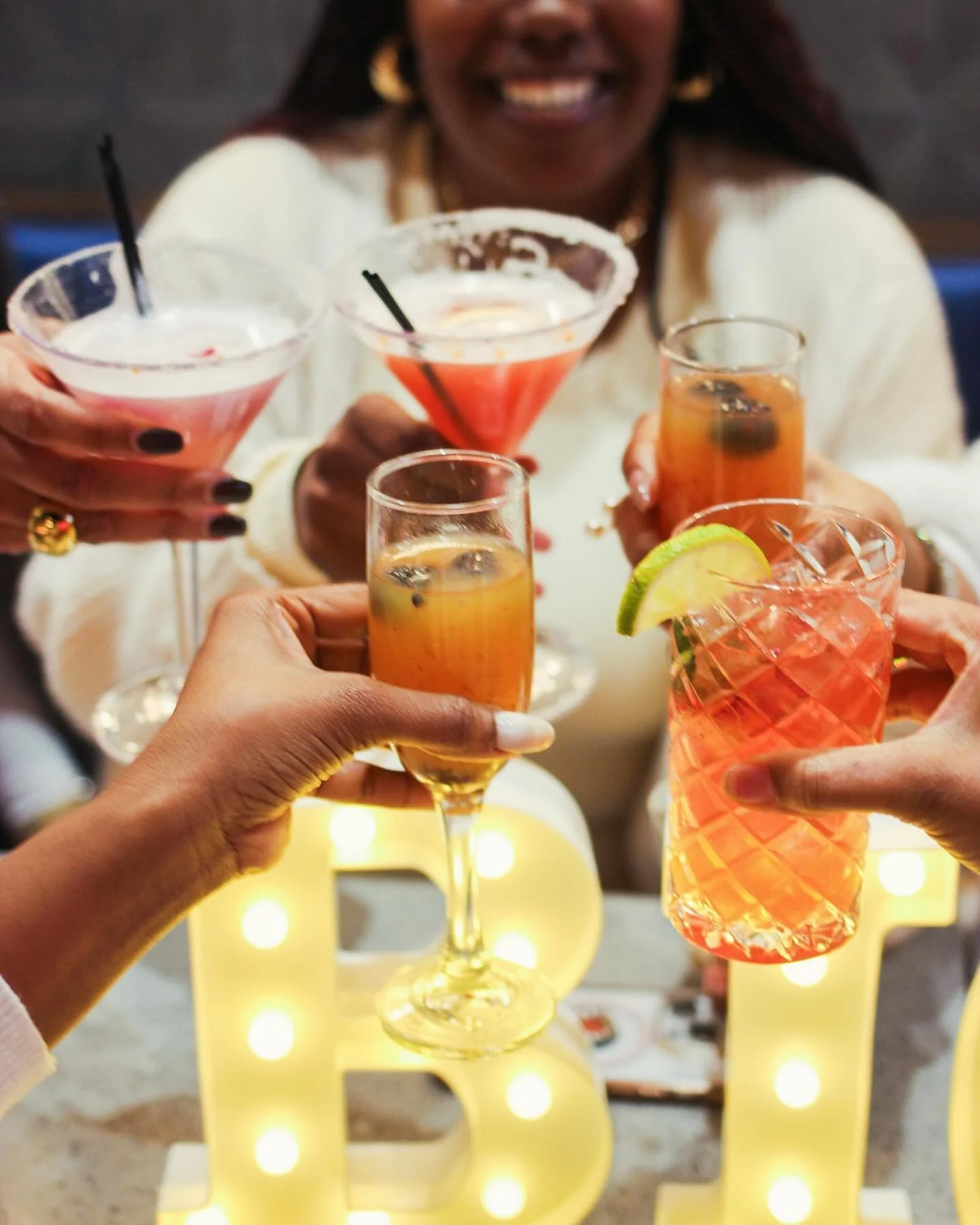 A group of people holding various cocktails and toasting in celebration with a lit letter 'B' decoration in the background.