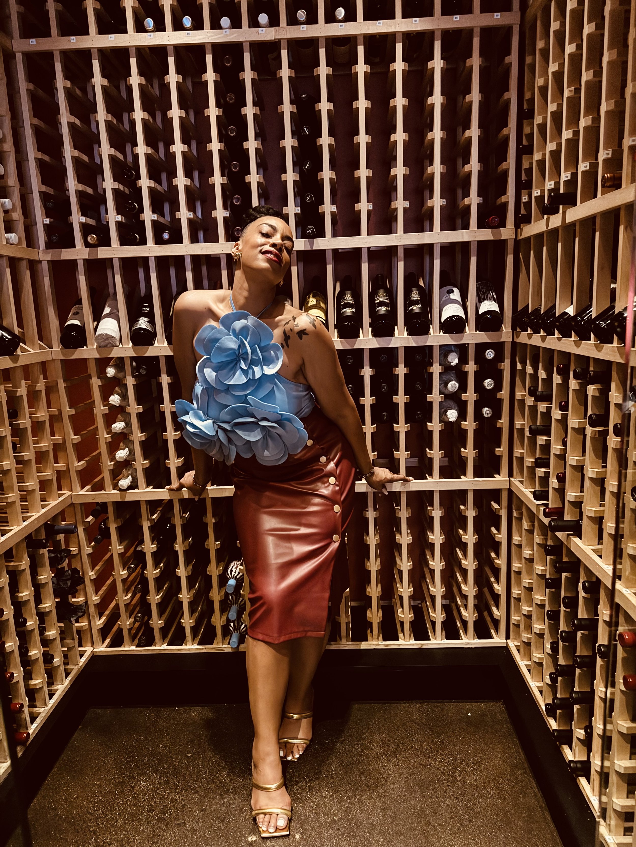 Person smiling, wearing a blue floral dress with birds tattooed on their shoulder, standing indoors near shelves with bottles.
