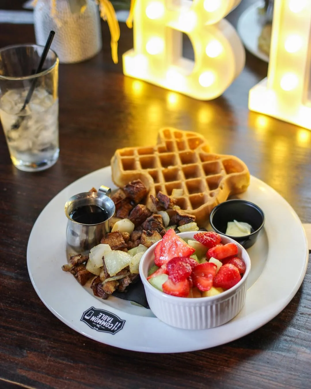 Plated breakfast with a waffle, diced potatoes, syrup, fruit bowl with strawberries, cucumbers, and melon, and a drink with ice on a wooden table.